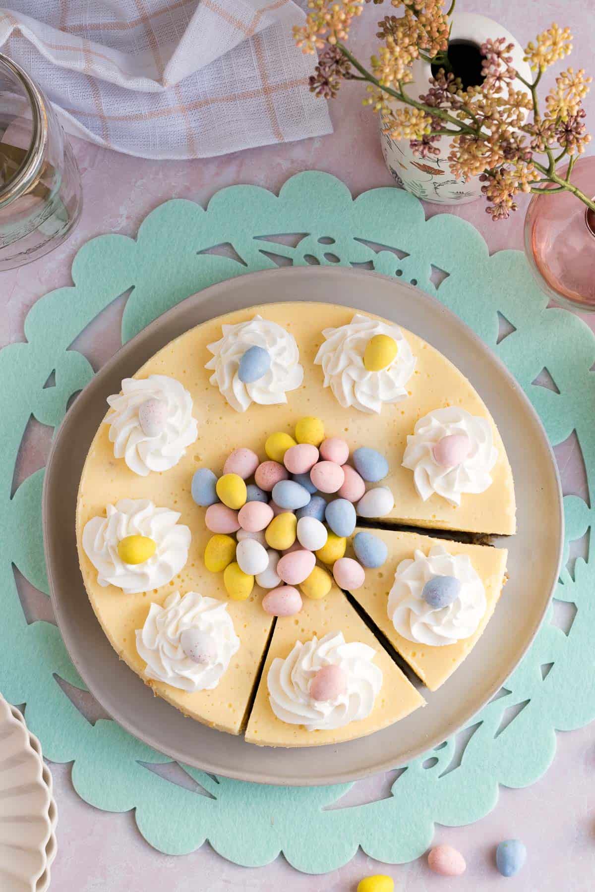 Top view of an Easter egg cheesecake with whipped cream and mini eggs piled on top and 2 slices cut sitting on a green Easter placemat