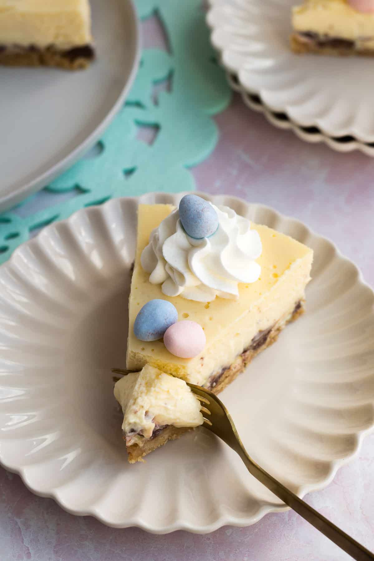 a cream scalloped plate with a slice of easter egg cheesecake with a gold fork cutting into it