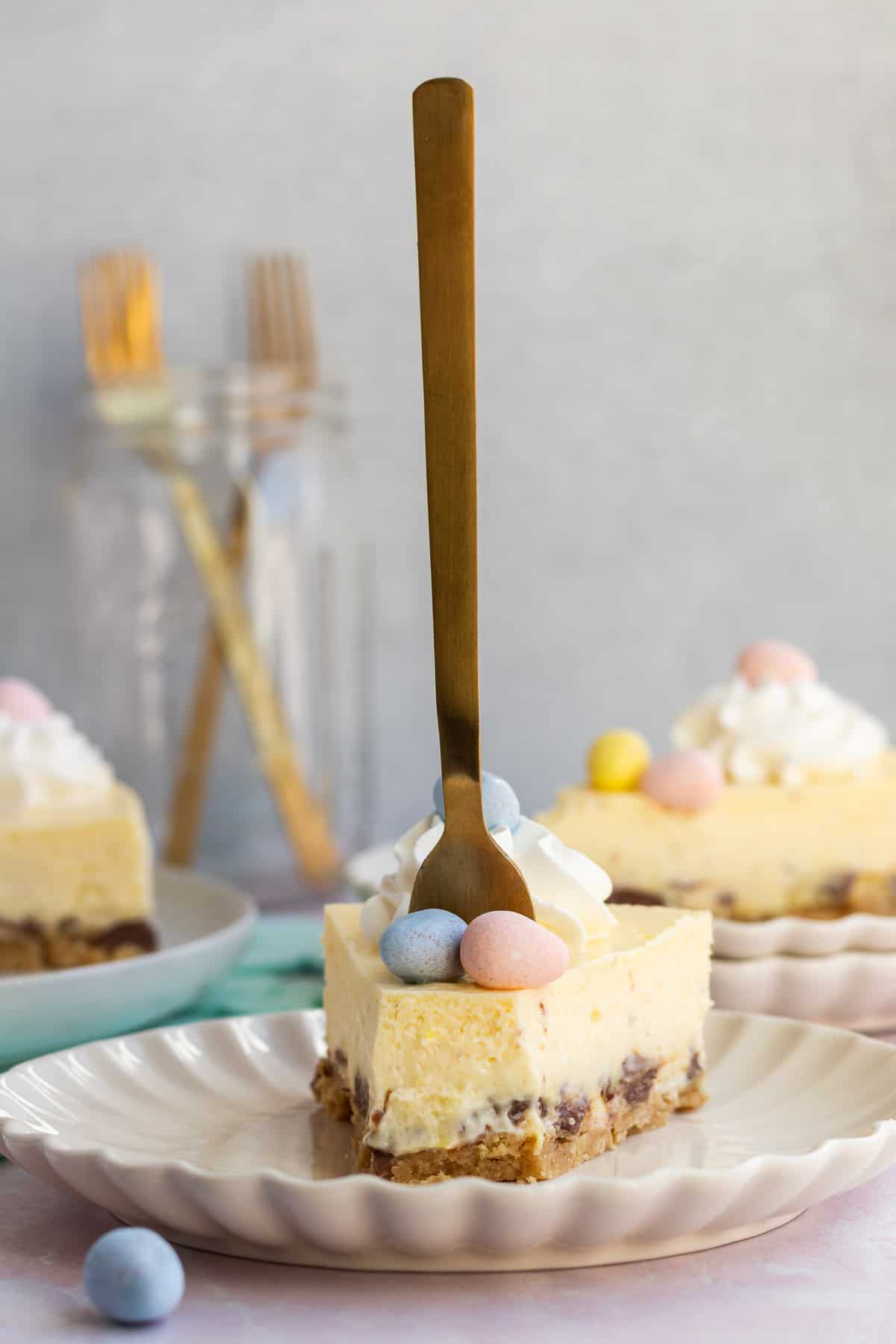 a slice of Easter egg cheesecake on a plate with a gold fork sticking up straight in it and other slices in the background