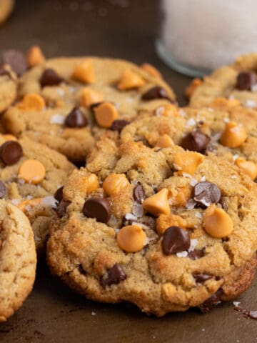 a pile of chocolate chip butterscotch cookies on a cookie sheet with a glass jar of sea salt