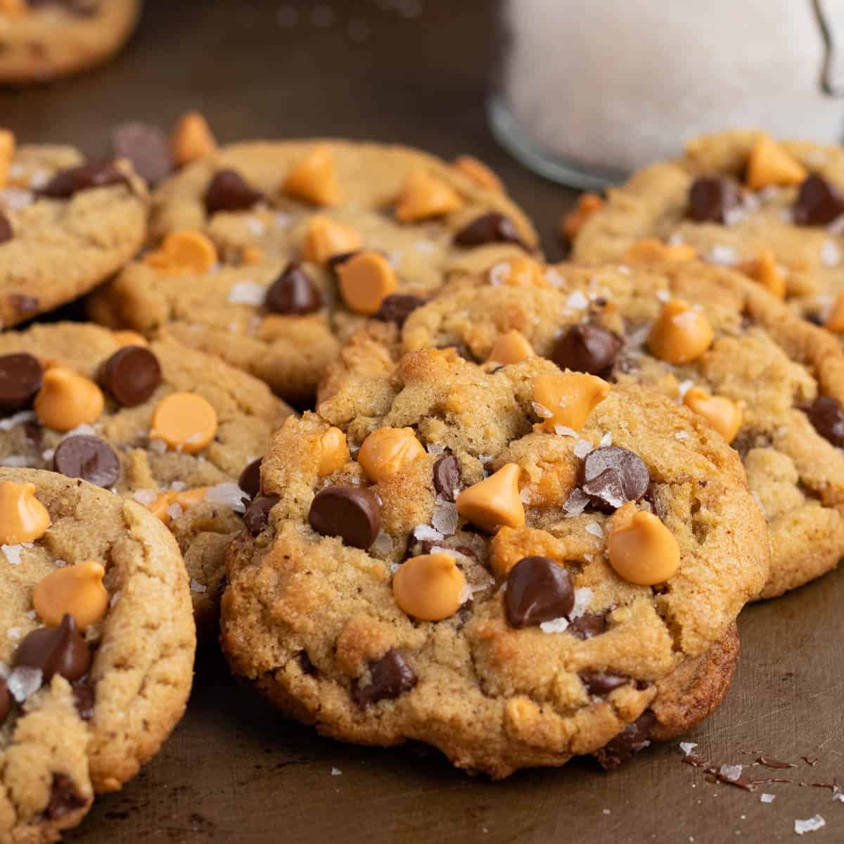 a pile of chocolate chip butterscotch cookies on a cookie sheet with a glass jar of sea salt 