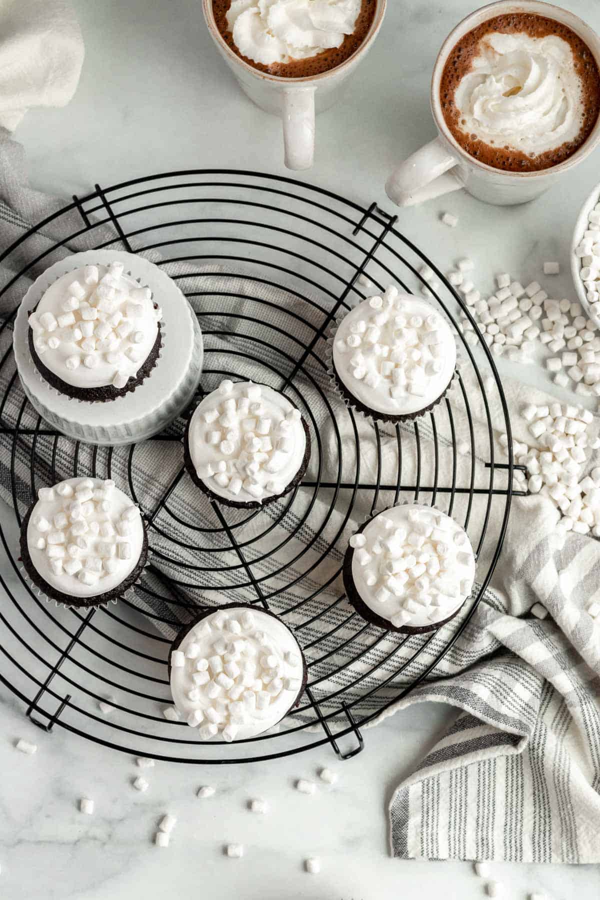 hot chocolate cupcakes on a wire cooling rack with 2 mugs of hot chocolate a mini marshmallows scattered around