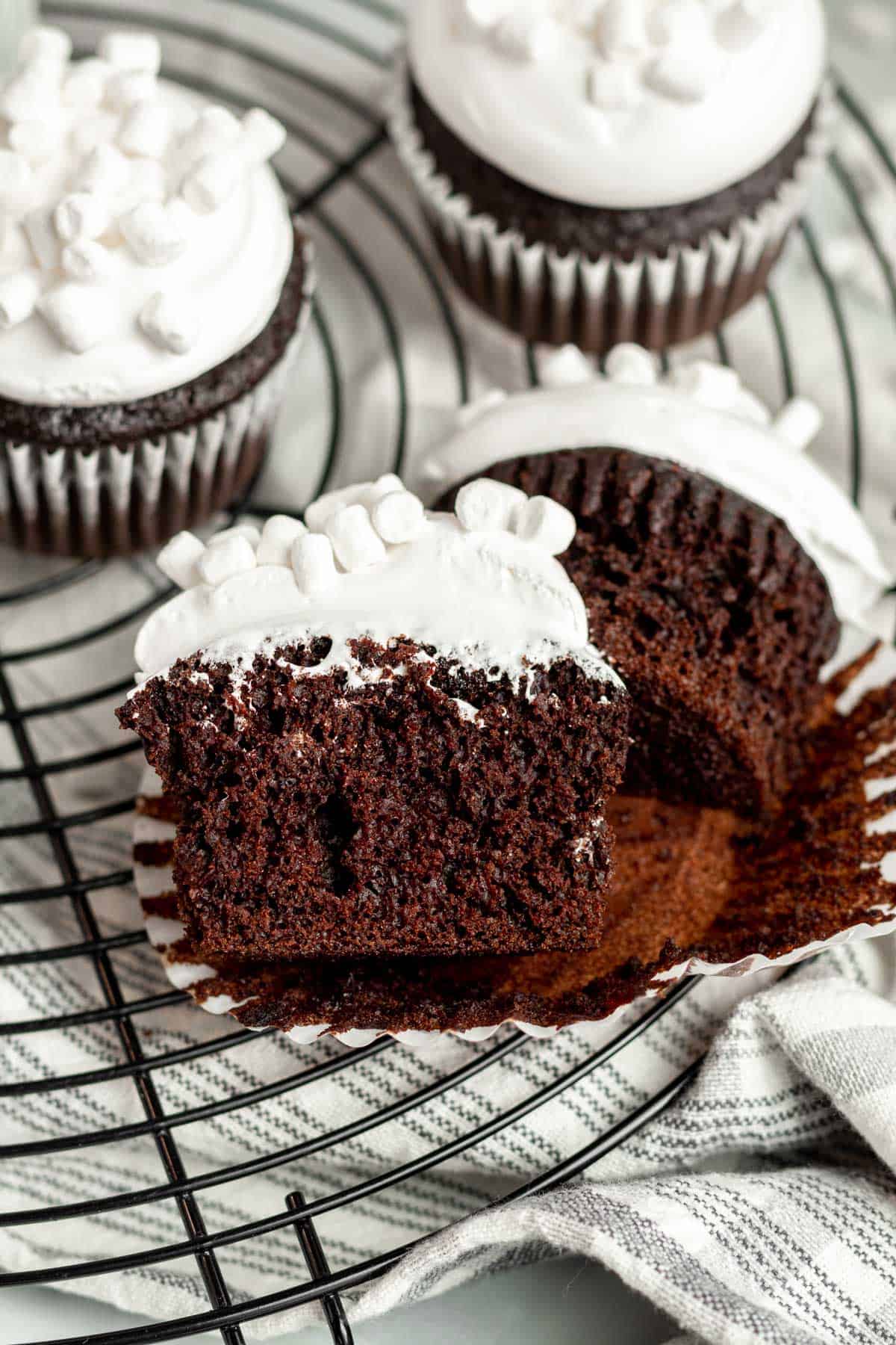 a hot chocolate cupcake with marshmallow frosting and mini marshmallows sliced open and sitting on top of a black wire cooling rack