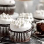 hot chocolate cupcakes topped with marshmallow buttercream and mini marshmallows sitting on a black cooling rack