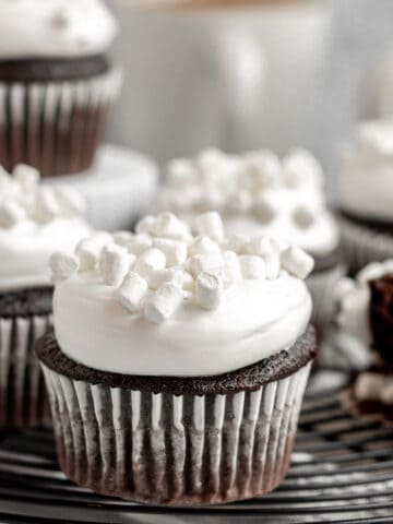 hot chocolate cupcakes topped with marshmallow buttercream and mini marshmallows sitting on a black cooling rack