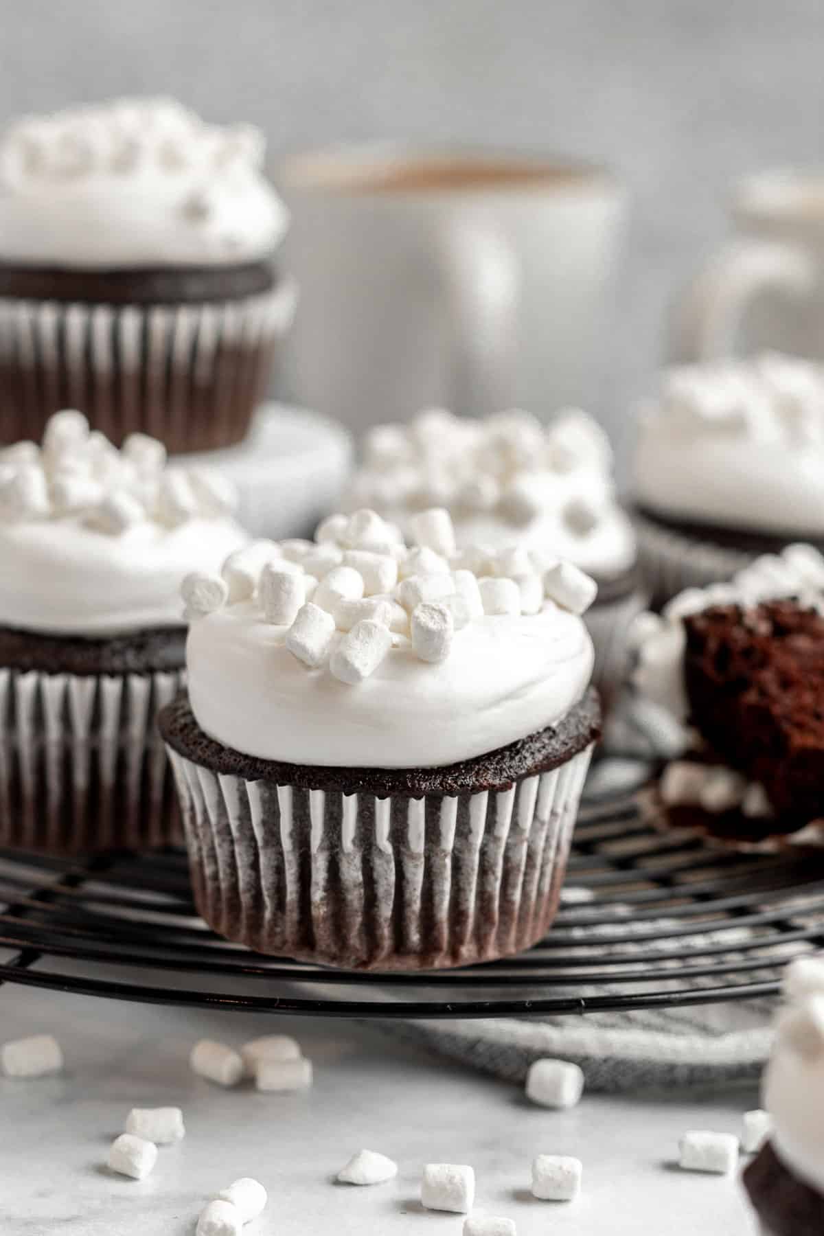 hot chocolate cupcakes topped with marshmallow buttercream and mini marshmallows sitting on a black cooling rack