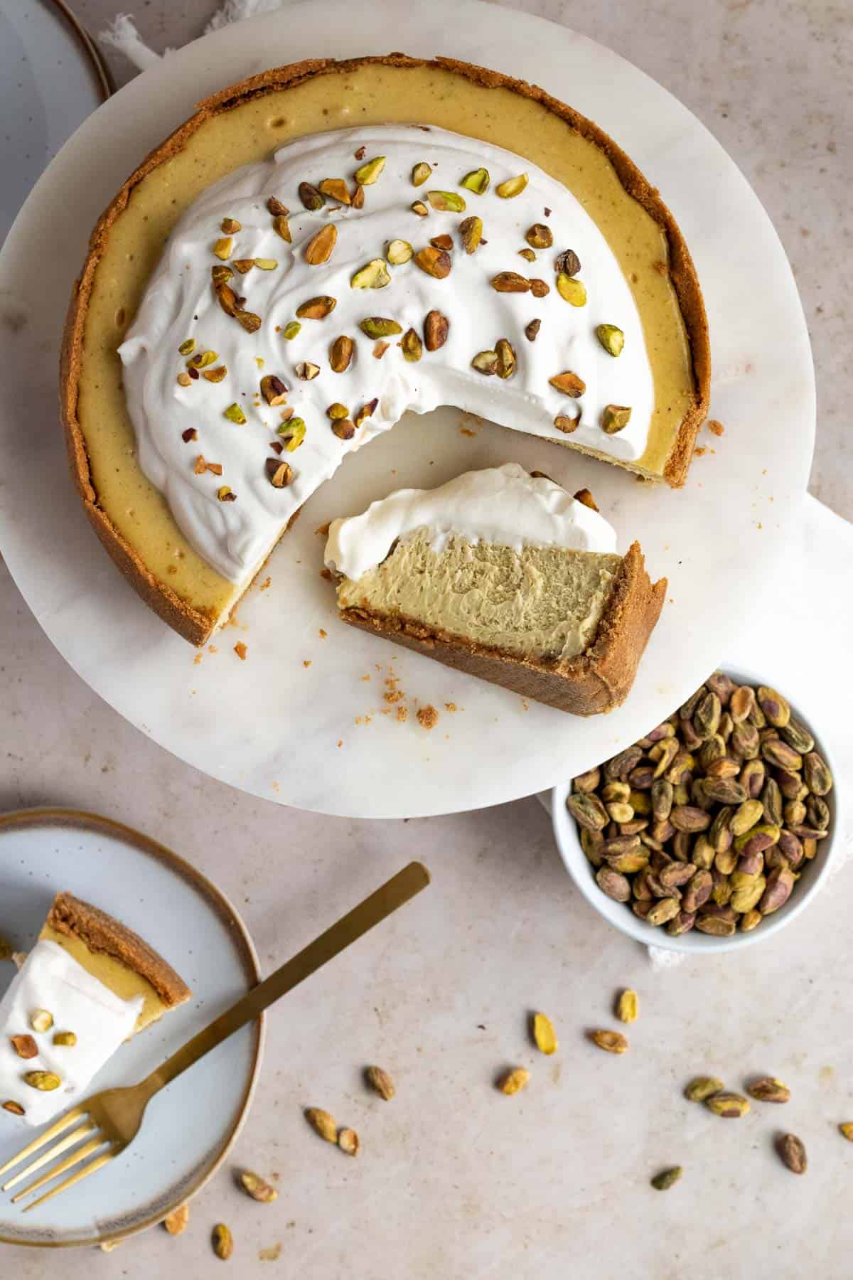 top view of a pistachio cheesecake on a marble cake stand with a slice on a plate and a bowl of pistachios nearby