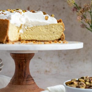 pistachio cheesecake on a wood and marble cake stand with a bowl of pistachios next to it