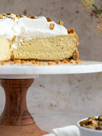 pistachio cheesecake on a wood and marble cake stand with a bowl of pistachios next to it