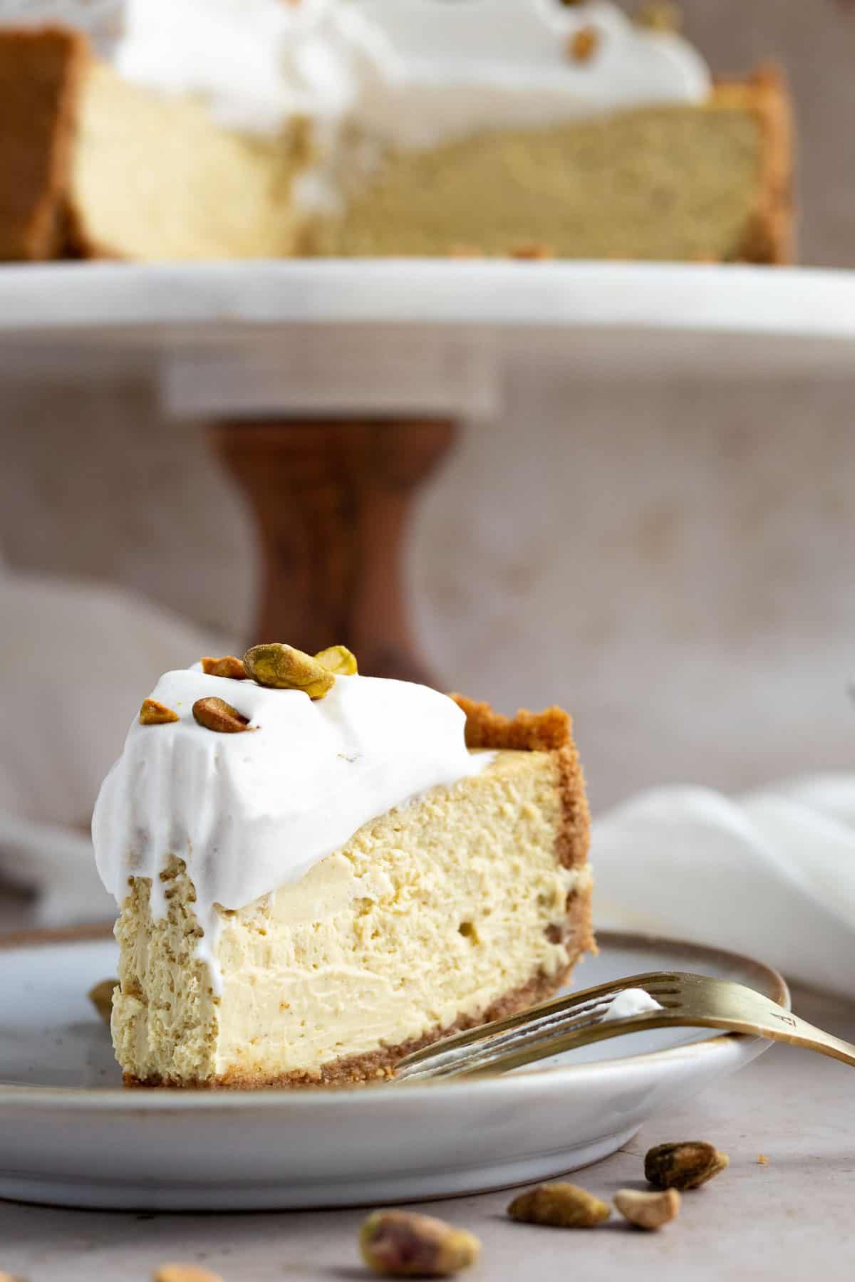 pistachio cheesecake slice on a plate with the whole cheesecake on a cake stand behind it