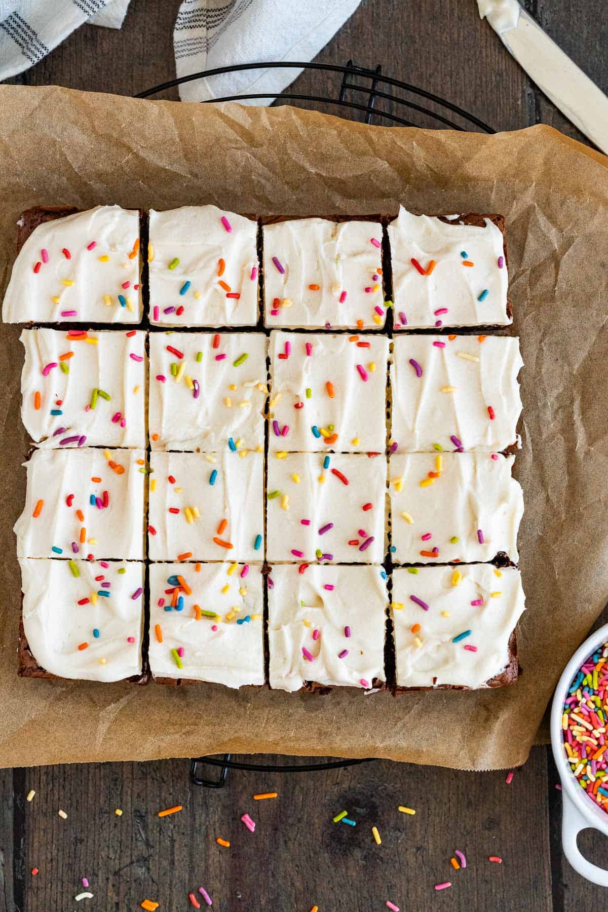 brownies with cream cheese frosting and sprinkles, sliced into a four by four grid and sitting on top of brown parchment paper