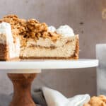 cinnamon toast crunch cheesecake on a wood and marble cake stand with slices cut out to show the inside of the cheesecake