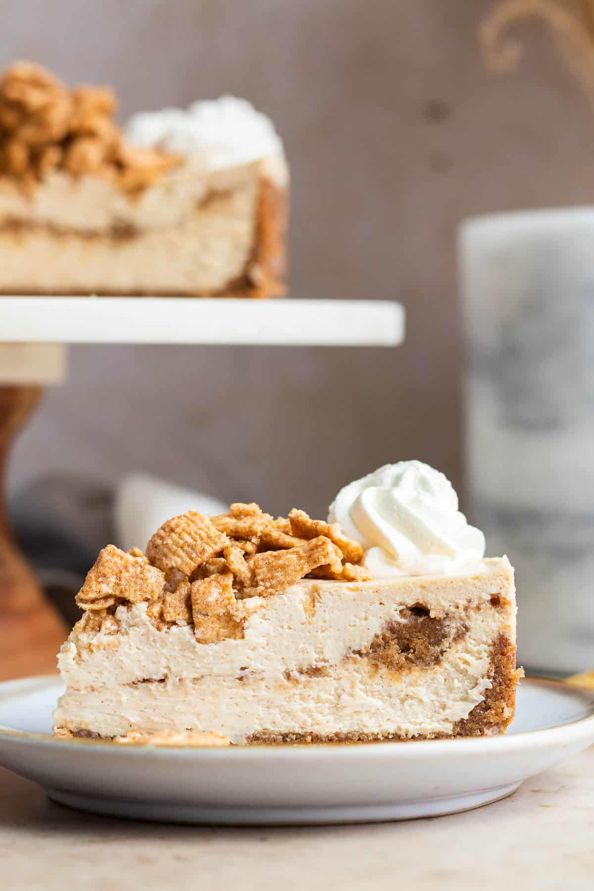 a slice of cinnamon toast crunch cheesecake on a white plate with the rest of the cheesecake in the background on a cake stand