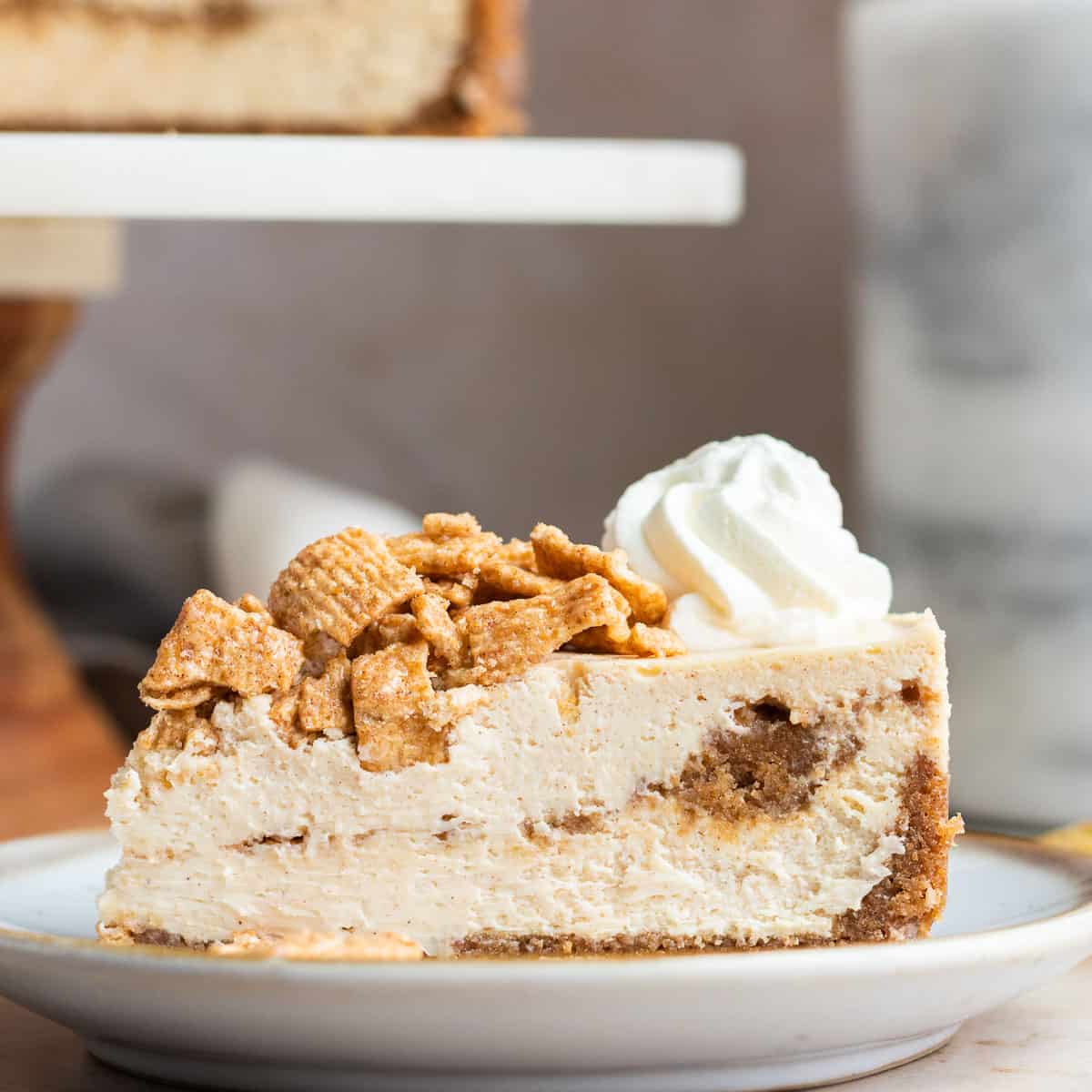 a slice of cinnamon toast crunch cheesecake on a white plate with the rest of the cheesecake in the background on a cake stand