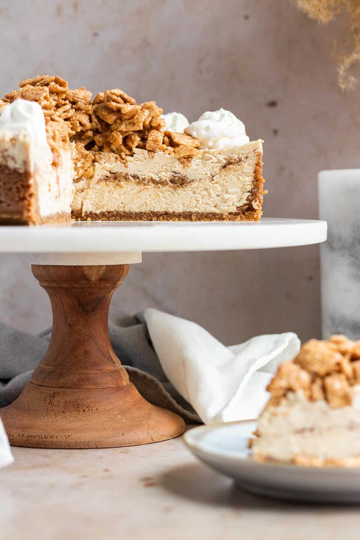 cinnamon toast crunch cheesecake on a wood and marble cake stand with slices cut out to show the inside of the cheesecake