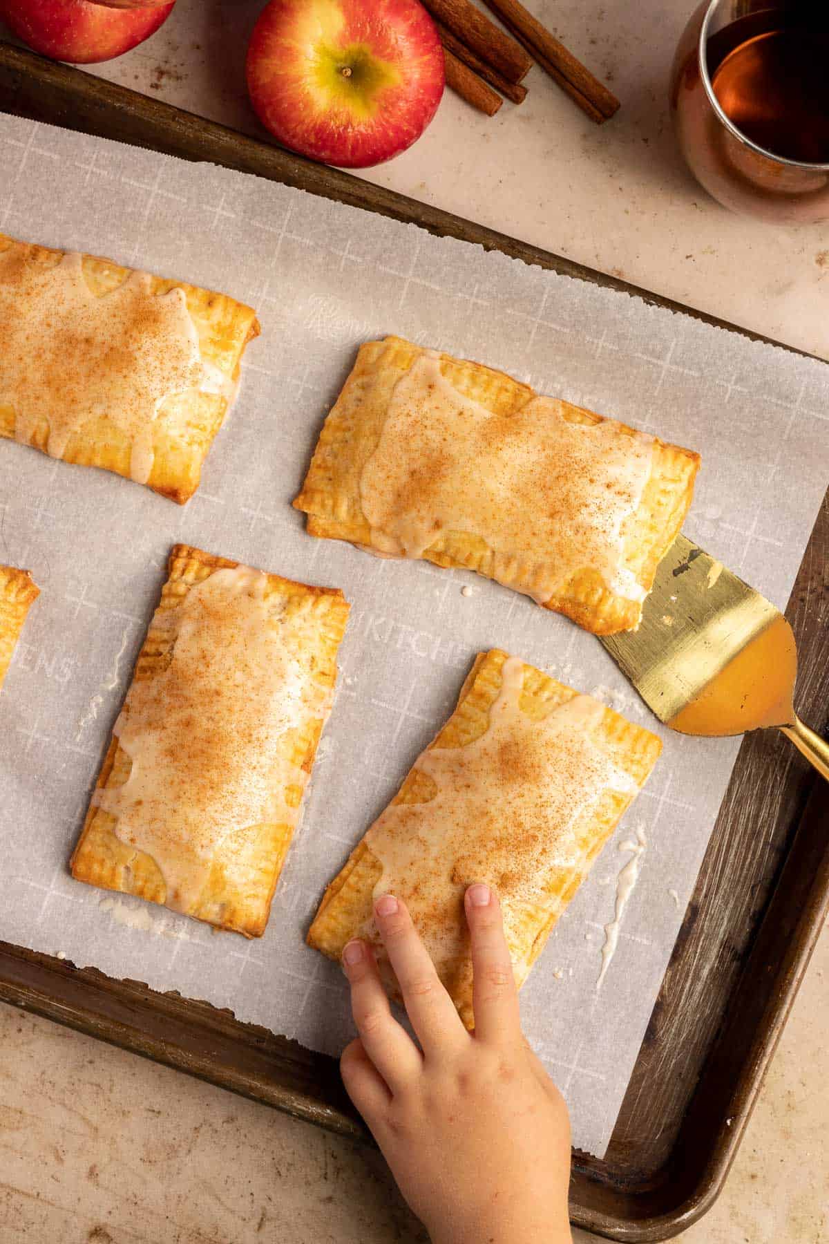 cinnamon apple pop tarts on a baking sheet with a child's hand grabbing one from the sheet