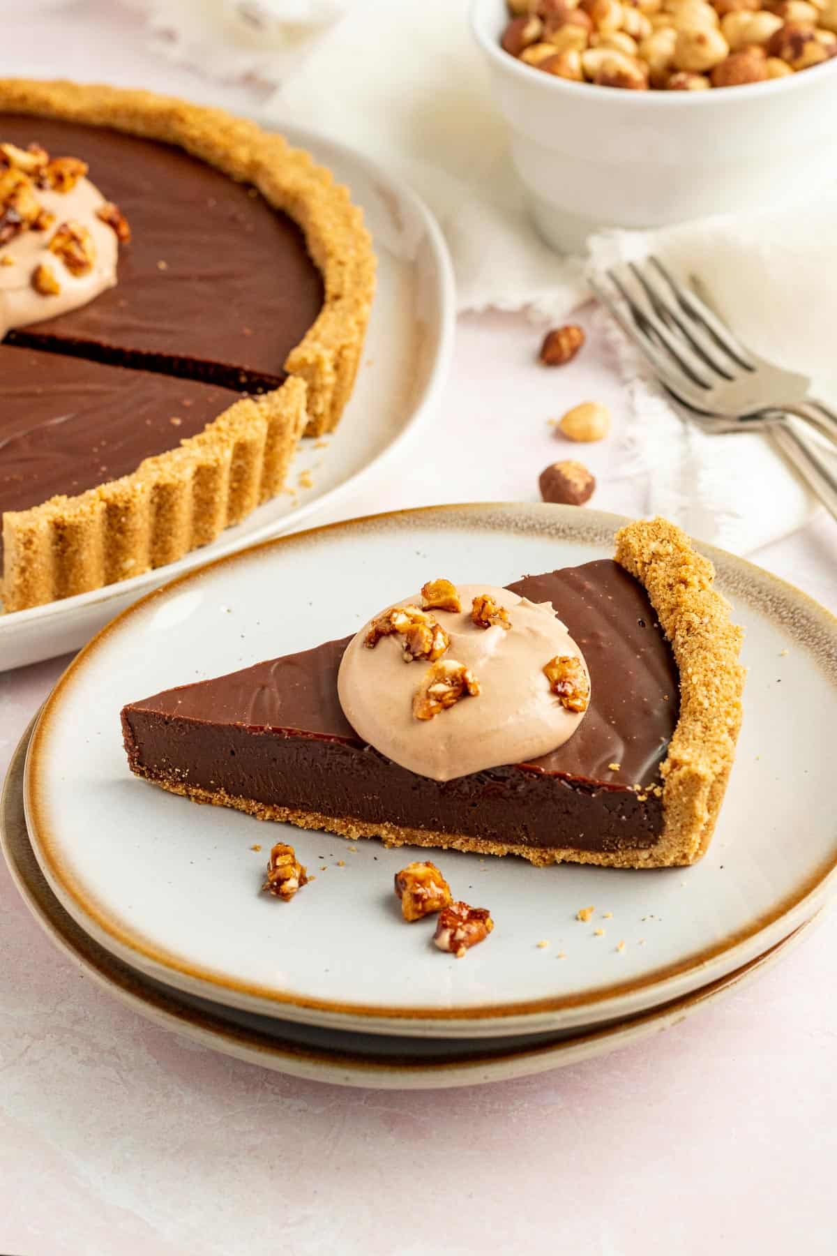 a slice of a nutella tart on a round white plate with a bowl of hazelnuts in the background and the whole tart in the background