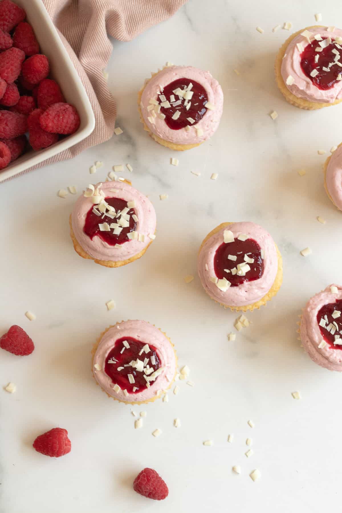 top view of white chocolate raspberry cupcakes with a container of raspberries on a marble background