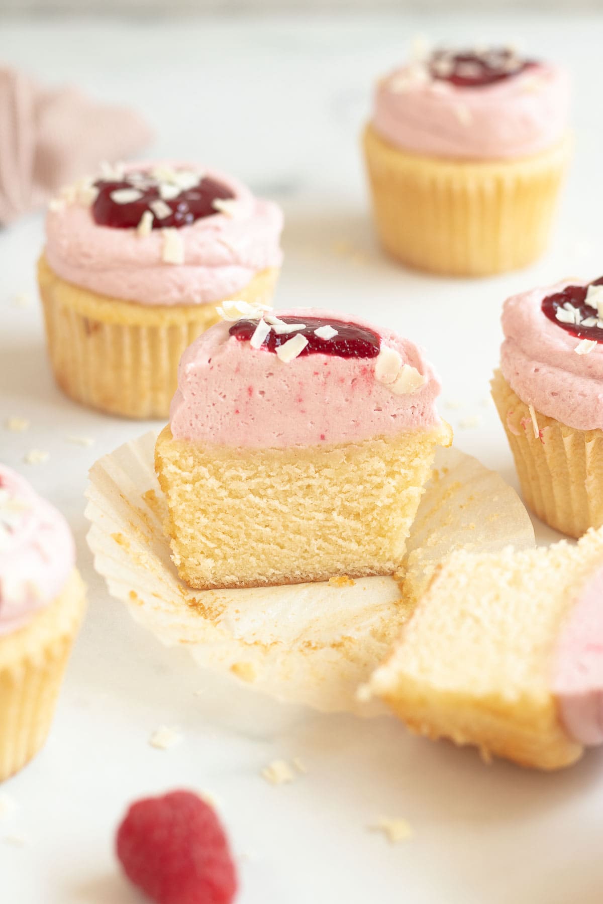 white chocolate raspberry cupcake with raspberry white chocolate buttercream and a dollop of raspberry sauce on top cut in half and laying on a cupcake wrapper