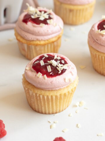 white chocolate cupcake in a white cupcake wrapper with pink frosting and raspberry sauce and shaved white chocolate on top