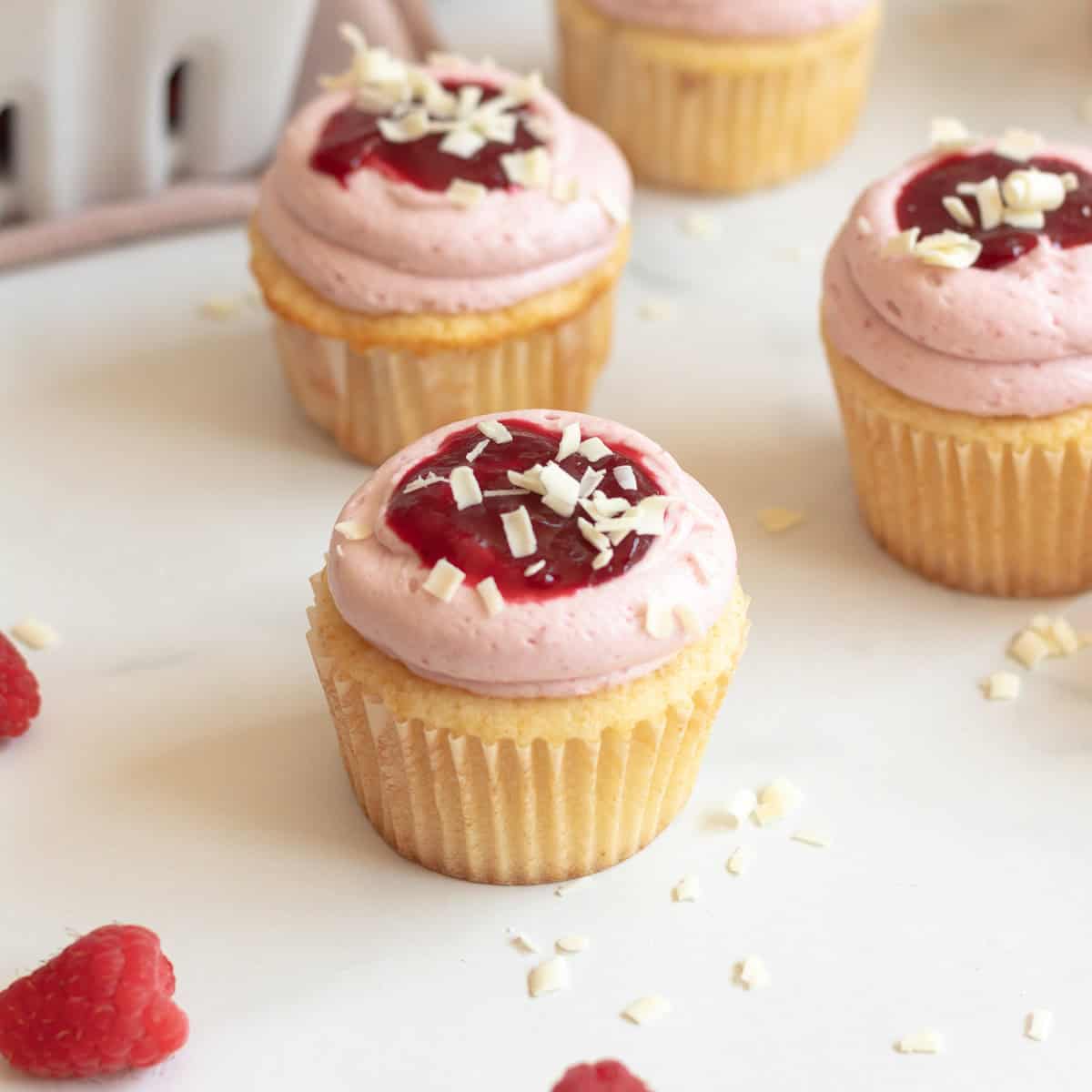 white chocolate cupcake in a white cupcake wrapper with pink frosting and raspberry sauce and shaved white chocolate on top